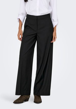Femme portant un pantalon noir à jambes larges, une chemise blanche à manches longues avec poignets retroussés, et des bottines marron, debout devant un fond uni.