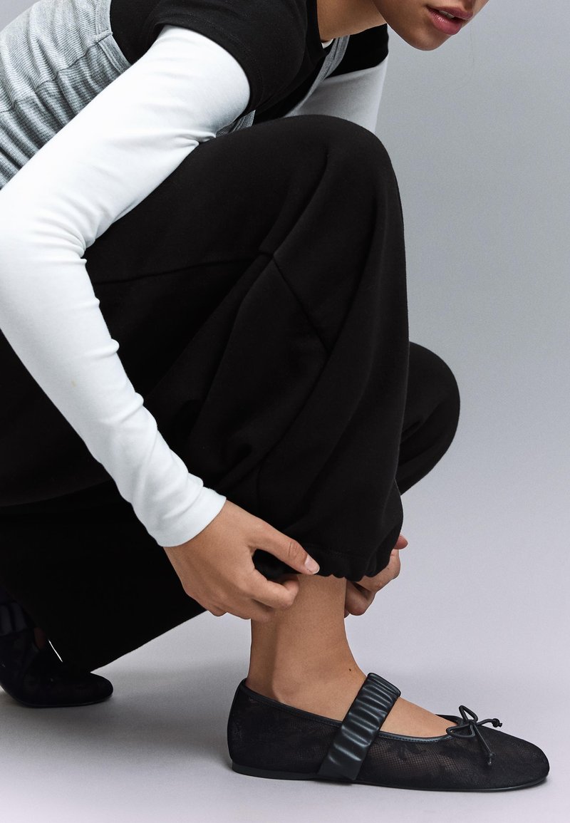 Personne accroupie, ajustant le poignet d'un pantalon noir, portant des ballerines noires avec une bride rembourrée, un haut blanc à manches longues sous des vêtements superposés gris et noirs.