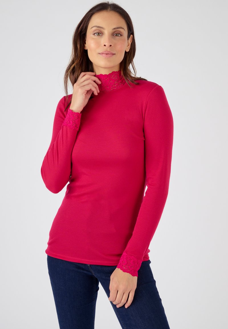Maglia attillata a maniche lunghe in tessuto rosa vivace con colletto alto in pizzo e dettagli in pizzo sui polsini, abbinata a jeans scuri.