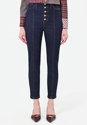 Frau trägt dunkelblaue, hochtaillierte Skinny-Jeans mit Knopfleiste, kombiniert mit bordeauxroten spitzen High Heels und einem gemusterten Langarmshirt.