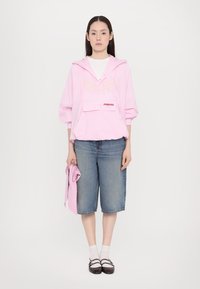 Rosa karierte Kapuzenjacke mit Fronttasche, kombiniert mit blauen Denim-Shorts. Das Model hält ein passendes pinkes Tuch, trägt weiße Socken und schwarze Schuhe.