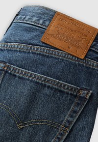 Jeans de mezclilla en azul oscuro con detalles de costura visibles. Presentan una etiqueta de cuero marrón con un logotipo en relieve y un diseño clásico de bolsillo.