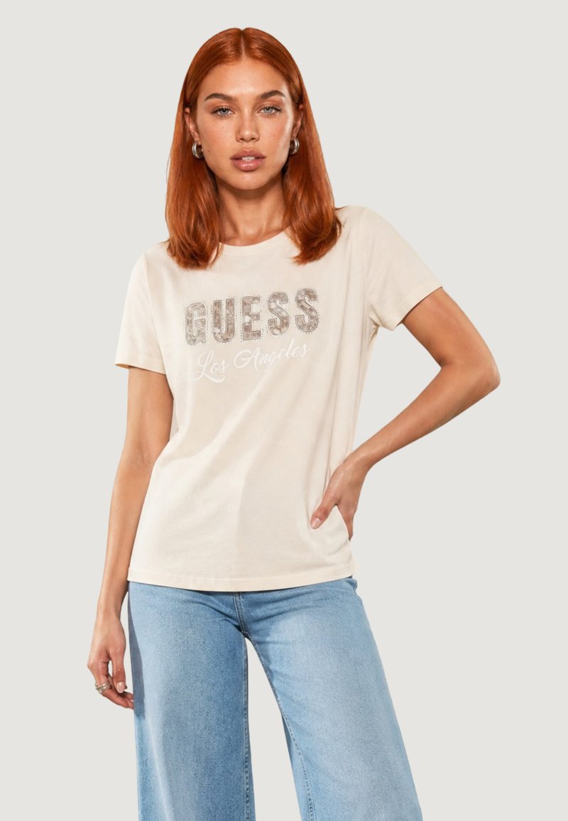 Femme aux cheveux roux portant un t-shirt beige Guess Los Angeles et un jean bleu clair, debout avec une main sur la hanche.