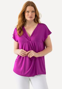 Fuchsiová krátká blůza s hlubokým výstřihem do V a zkrouceným předním detailem; lehký materiál s hladkou strukturou, zkombinováno s bílými kalhotami.