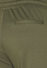 Olijfgroene broek met een gladde textuur, voorzien van een elastische tailleband en een enkele achterzak met een bijpassende stofaccent.