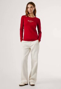 Raudona ilgomis rankovėmis marškinėliai su balta emblema, derinami su platėjančiomis kreminėmis kelnėmis ir šviesiais sportbačiais. Paprastas dizainas, lygus tekstūra.