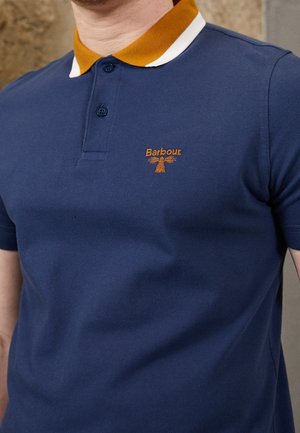 Mężczyzna w granatowej koszulce polo z kontrastującym musztardowo-białym kołnierzem, z pomarańczowym logo Barbour na lewym biuście.