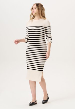 Femme portant une robe tricotée crème avec des rayures horizontales noires et des manches longues, marchant avec des chaussures slingback noires à bout pointu sur fond blanc.
