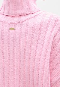 Gros plan d’un pull à col roulé en maille côtelée rose avec une petite étiquette rectangulaire dorée de la marque dans le dos, près de l’encolure.