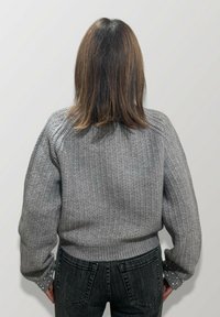Persona con capelli castano scuro alla lunghezza delle spalle, che indossa un maglione grigio a maglia e jeans scuri, in piedi contro uno sfondo chiaro e uniforme, visto da dietro.