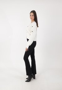 Witte gebreide cropped cardigan met zwarte hartpatronen, gecombineerd met zwarte uitlopers en zwarte haklaarzen. Effen witte achtergrond.