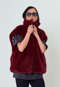 Burgundi spalvos dirbtinio kailio liemenė su aukštu apykakle, užtrauktuku ir trumpomis rankovėmis, derinama su tamsiai pilku marškinėliu ir juodais dryžuotais kelnių.