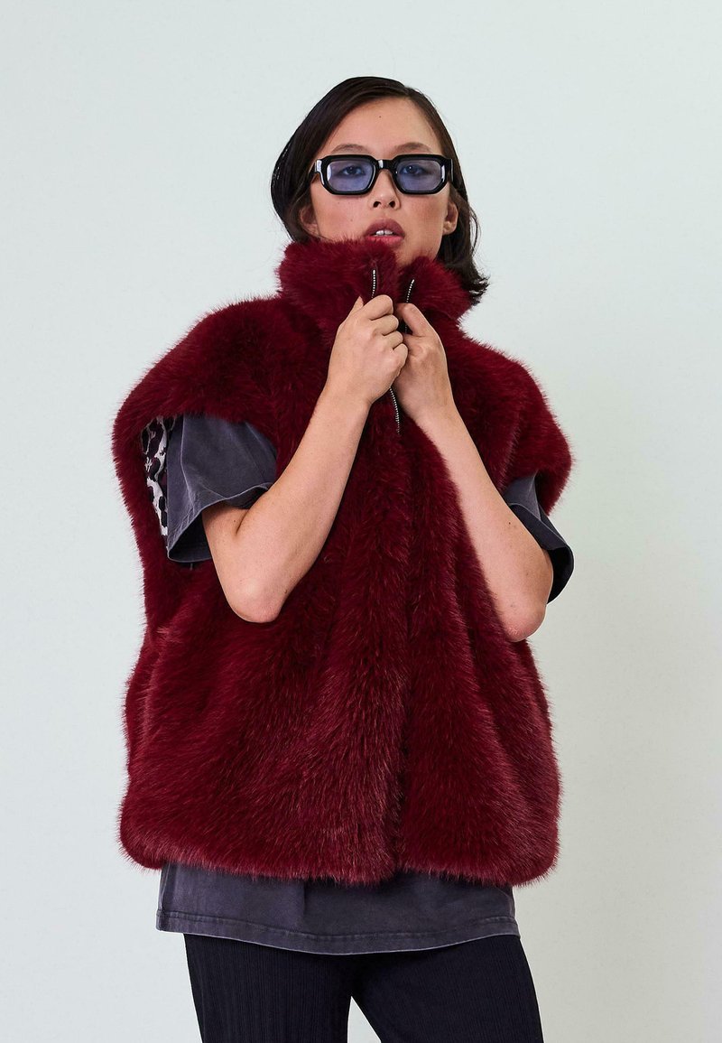 Burgundi spalvos dirbtinio kailio liemenė su aukštu apykakle, užtrauktuku ir trumpomis rankovėmis, derinama su tamsiai pilku marškinėliu ir juodais dryžuotais kelnių.