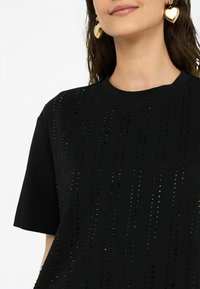 Schwarzes T-Shirt mit kurzen Ärmeln, das vertikale Reihen kleiner, glänzender schwarzer Verzierungen zeigt. Weicher Stoff mit Rundhalsausschnitt.
