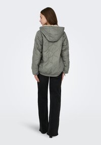 Topattu oliivinvihreä takki hupulla, jossa on tikattu pyöreä kuvio. Yhdistetty mustiin leveälahkeisiin housuihin, joissa on rento istuvuus.