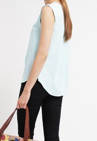 Blusa senza maniche azzurro chiaro con orlo arrotondato, abbinata a pantaloni neri. Il modello tiene una borsa a tracolla multicolore con manici marroni.