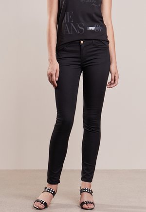 Vrouw die zwarte skinny jeans draagt, een zwart mouwloos topje met bedrukte tekst en zwarte sandalen met zilveren studs, staand op een neutrale vloer.