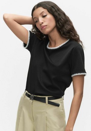 Kvinde med langt krøllet hår iført en sort T-shirt med hvide kanter, beige bukser med høj talje og et sort bælte, poserer med den ene arm løftet.