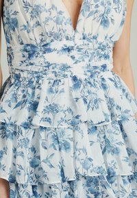Vestido floral con un patrón azul y blanco, capas con volantes, escote en V profundo y una tela ligera y texturizada.