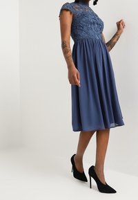 Kvinna klädd i en knälång blå klänning med spetsöverdel och svarta högklackade skor, stående mot en ljus och enkel bakgrund.