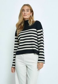 Schwarz-beige gestreifter Pullover mit hochgestelltem Kragen, langen Ärmeln und lockerer Passform, kombiniert mit hellen, maßgeschneiderten Hosen.