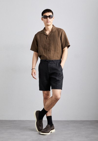 Cămașă scurtă din in maro, cu mâneci scurte și nasturi, pantaloni scurți negri și pantofi loafers din nubuc închis, cu tălpi albe. Modelul poartă ochelari de soare negri.