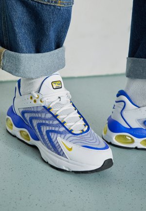 Baskets blanches, bleues et jaunes portées avec des chaussettes blanches et un jean bleu retroussé, debout sur un sol gris clair tacheté.