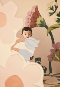 Spalvingos išpjaustytos gėlių formos iš minkštų medžiagų apsupa jauną vaiką, kuris laiko šviesiai mėlyną gėlių gabalėlį prieš raštuotą grindų dangą.