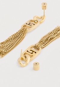 Náušnice z kovu so zlatým odtieňom v tvare reťazí s viacerými jemnými reťazcami a obdĺžnikovými značkovými príveskami, jeden voľný podklad viditeľný na bielom pozadí.