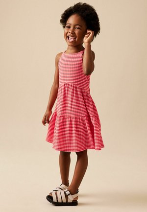 Smiling child with curly hair stands wearing a red checkered sleeveless dress and white sandals against a beige background.