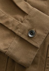 Tan fabric shirt cuff featuring double stitching, a rounded design, and a silver metal button with engraved text. Smooth texture.