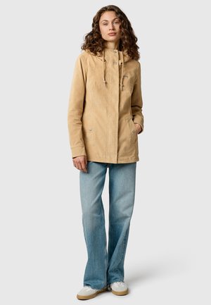 Donna con capelli ricci che indossa una giacca beige con cappuccio, jeans larghi color azzurro chiaro e scarpe da ginnastica bianche, in piedi con una mano in tasca.