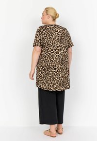 Leopardenmuster, Tunika mit kurzen Ärmeln; beiger Hintergrund mit braunen Flecken. Kombiniert mit schwarzen, weit geschnittenen Hosen und flachen Riemchensandalen.
