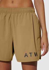 Beige shorts met een elastische tailleband, zijzakken en een zwarte "ATW" print op de linkeronderkant. Gladde textuur en een relaxte pasvorm.