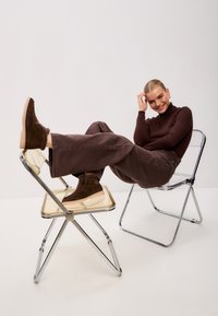 Bottes de cheville en daim marron avec une finition texturée, dotées d'une semelle beige clair, associées à un pantalon ample marron foncé sur une chaise pliante en chrome.