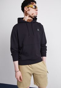 Black hoodie with a front pocket and drawstring, featuring a small logo. Paired with beige shorts, model wearing sunglasses.
