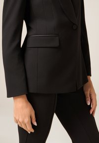 Czarna, dopasowana marynarka na jedno zapięcie, z wyraźnie zarysowanymi ramionami i gładkim, eleganckim materiałem. Po lewej stronie widoczna boczna kieszeń.