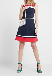 Marindress med röd kant och vita ränder, ärmlös design, matchad med en liten vit handväska och gröna klacksandaler.