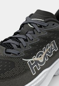 Chaussure de sport noire avec une tige en mesh et textile, lacets gris et un logo HOKA bien en vue. Semelle texturée et éléments de design épurés.