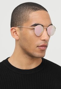 Ochelari rotunzi cu lentile roz și rame metalice subțiri, purtați de o persoană într-un pulover negru cu model băț. Design curat, hardware minim.