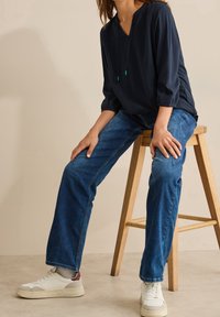 Blusa blu navy, ampia e comoda, con scollo a V e cordini verdi, abbinata a jeans di denim blu e sneakers bianche, seduta su uno sgabello di legno.