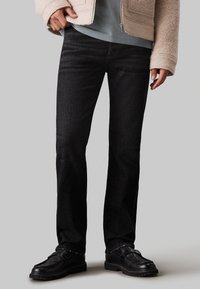 Jean en denim noir avec une coupe droite, un léger délavage et une texture subtile, associé à une veste en polaire beige et des mocassins noirs.