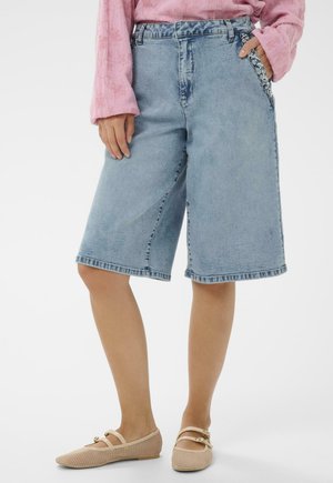 Osoba nosi svetlomodré rifľové šortky nad kolená s ozdobenými vreckami, ružový dlhý rukáv a béžové sieťované balerínky.