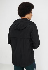 Svart lättviktig jacka med huva, med en texturerad yta och lös passform. Designen inkluderar en bakre ventil för luftflöde.