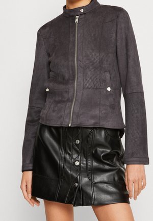 Femme portant une veste zippée gris foncé perforée et une mini-jupe en cuir noire à boutons, debout, les mains détendues le long du corps.