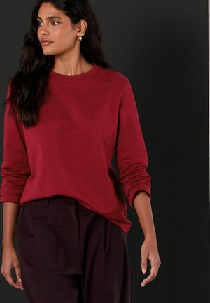 Rotes Langarmshirt mit rundem Ausschnitt, glattem Stoff und Bündchenärmeln, kombiniert mit dunklen, plissierten Hosen.