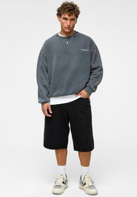 Grijze oversized sweatshirt met verlaagde schouders, zwarte denim shorts, een wit T-shirt eronder, en beige sneakers met zwarte accenten.