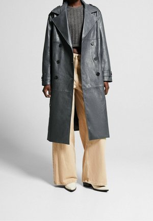 Person iført en lang mørkegrå læder trenchcoat, grå kort strikket sweater, beige bukser med vide ben og hvide spidse støvler.