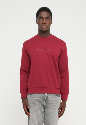Rød sweatshirt med rund hals og ribbede manchetter, prydet med et gråt trykt logo "Armani Exchange" på tværs af brystet. Bæres med grå jeans.