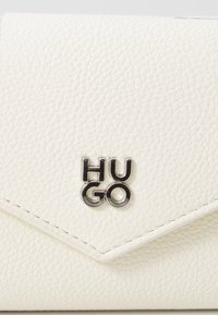 Portefeuille en cuir blanc texturé avec un logo "HUGO" argenté. Présente un design à rabat pointu et des coutures visibles. Compact et minimaliste.